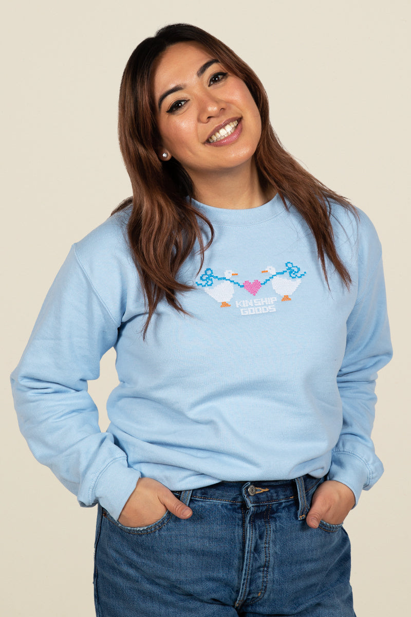 Kin Ship Goods blue sweatshirt with cross stitch art of two geese holding a heart between them and 'Kin Ship Goods' text below.